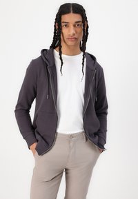 Sudadera con cremallera en algodón gris oscuro, con capucha forrada, bolsillos frontales y cordones ajustables, combinada con una camiseta blanca y pantalones beige.