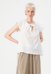 Blusa bianca senza maniche con spalle arricciate, scollo legato e righe verticali. Tessuto leggero, vestibilità ampia, abbinata a una gonna plissettata color beige.