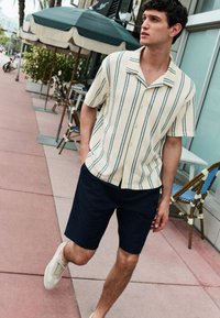 Short-sleeve, beige striped button-up shirt with dark green lines, paired with navy shorts and light sneakers, set against a sidewalk café scene.