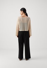 Haut en tricot ouvert de couleur crème à manches longues sur un pantalon large noir. Le tissu présente une texture légère et aérée ainsi qu'un motif géométrique.