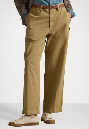 Homme portant un pantalon beige à jambes larges, une ceinture en cuir marron, une chemise en denim bleu, une veste marron et des baskets beiges à lacets, debout les mains dans les poches.