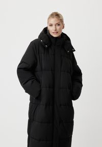 Femme aux cheveux blonds portant un long manteau noir rembourré avec une capuche, les mains dans les poches, regardant la caméra sur un fond blanc.
