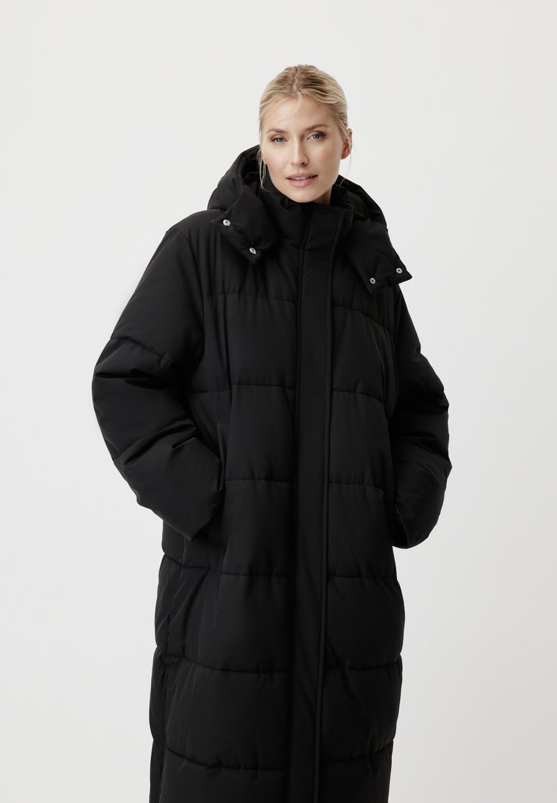 Femme aux cheveux blonds portant un long manteau noir rembourré avec une capuche, les mains dans les poches, regardant la caméra sur un fond blanc.