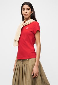 Κόκκινο βαμβακερό μπλουζάκι με κοντά μανίκια, με μικρό κεντητό λογότυπο. Μπεζ πλεκτό κασκόλ κρέμεται γύρω από τον λαιμό. Πλισέ χακί φούστα.