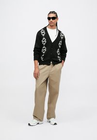 Homme avec des cheveux tressés et des lunettes de soleil portant un cardigan noir avec des motifs de serpent, une chemise blanche, un pantalon beige et des baskets blanches devant un fond uni.