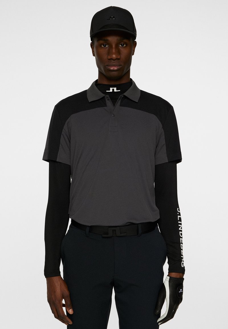 Svart keps och långärmad skjorta under en kortärmad polo. Polon är mörkgrå med svart mönster; har logotypdetaljer och handskar.