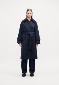 Cappotto di lana blu scuro con colletto, chiusura a bottoni e cintura. Presenta maniche ampie con dettagli a bottoni. Indossato sopra pantaloni di jeans scuri.