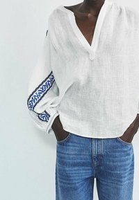 Witte linnen top met een V-hals en lange mouwen, voorzien van blauwe geborduurde patronen op de mouwen. Draag het met blauwe denim jeans.