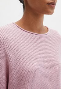 Femme portant un pull rose clair texturé en tricot avec un col arrondi, visible des épaules au menton sur un fond uni.