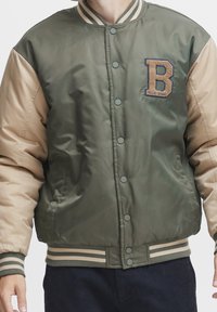 Groene en tan bomberjack met geribbelde kraag, knoopsluiting en geborduurde letter "B" op de linkerkant van de borst. Gestructureerde stof, gewatteerd ontwerp.