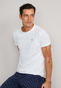 Bílý tričko s krátkým rukávem a kulatým výstřihem, na hrudi s malým černým emblémem. Nosené s námořnickými vzorovanými šortkami.