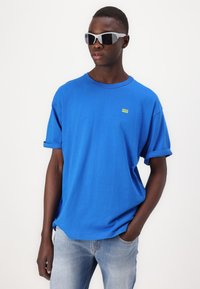 Camiseta azul de manga corta de algodón, con cuello redondo y un pequeño logo amarillo en el pecho. Llevada con unos jeans de un azul claro.