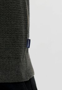 Pull en maille texturée gris foncé vu de côté avec une petite étiquette bleue marine affichant la marque "Jack & Jones" sur un fond clair uni.