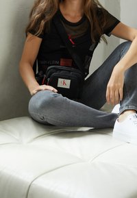 Černá křížová taška z látky s červenými zipy, se stříbrným logem CK. Modelka má na sobě průsvitný černý top a šedé džíny. Bílé tenisky.