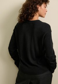 Maglione nero a maglia con scollatura e polsini a coste, caratterizzato da una texture liscia e una vestibilità rilassata. La schiena del modello è rivolta verso la macchina fotografica.