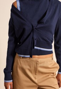 Cardigan lavorato a maglia blu navy con dettagli azzurri, dotato di scollo a V, chiusura con bottoni e polsini a costine, abbinato a pantaloni beige.