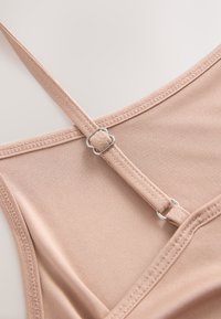 Hellbeige Kleidungsstück mit strukturiertem Stoff, das verstellbare Träger und ein silbernes Metallteil auf der Rückseite aufweist.