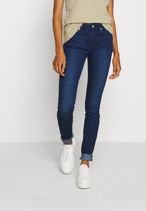 Dark blue skinny jeans feature a mid-rise waist, fitted silhouette, and cuffed hems, paired with a beige t-shirt and white sneakers.