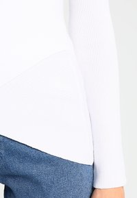 Haut blanc à manches longues côtelé avec un design asymétrique, associé à un jean denim bleu foncé avec une finition texturée.