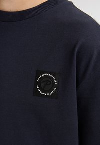 Felpa navy con patch rotondo nero e testo logo bianco. Collo a coste e texture di tessuto liscia. Design minimalista.