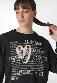 Svart sweatshirt med olika textdesign i vitt och ljusrosa, med en stor rosa hjärtform i mitten, tillverkad av bomull.