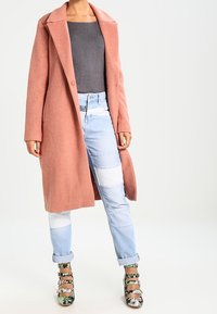 Peach-colored, long wool coat with a notched collar and button closure, paired with light blue, patchwork denim jeans and patterned heels.
