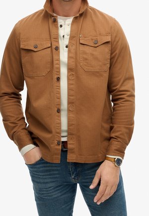 Homme portant une chemise marron boutonnée par-dessus un henley blanc et un jean bleu, avec une montre au poignet gauche et une main dans la poche.