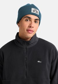 Jongeman draagt een teal gebreide beanie met het Jack Wolfskin-logo en een zwarte fleecejack met rits en Jack Wolfskin-patch.