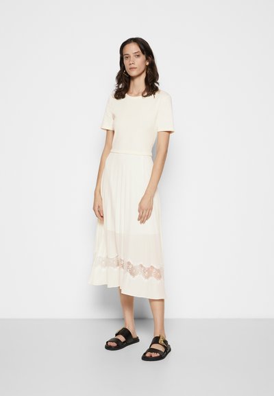 Robe crème à manches courtes avec une jupe plissée, ornée de détails en dentelle à l'ourlet. Associée à des sandales noires avec une semelle épaisse.