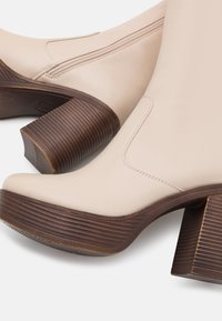 Bottes beige en cuir à cheville avec un bout arrondi, une fermeture éclair latérale et un talon bloc en bois marron avec des rainures horizontales.