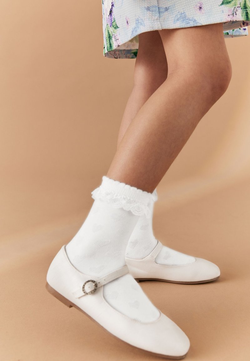 Enfant portant des chaussures blanches Mary Jane avec boucle et des chaussettes blanches bordées de dentelle, debout devant un fond beige.