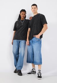 Tricouri supradimensionate cu imprimeu tie-dye negru, asortate cu blugi denim cu picioare largi de culoare albastru deschis. Modelul masculin poartă pantofi negri; modelul feminin poartă adidași.