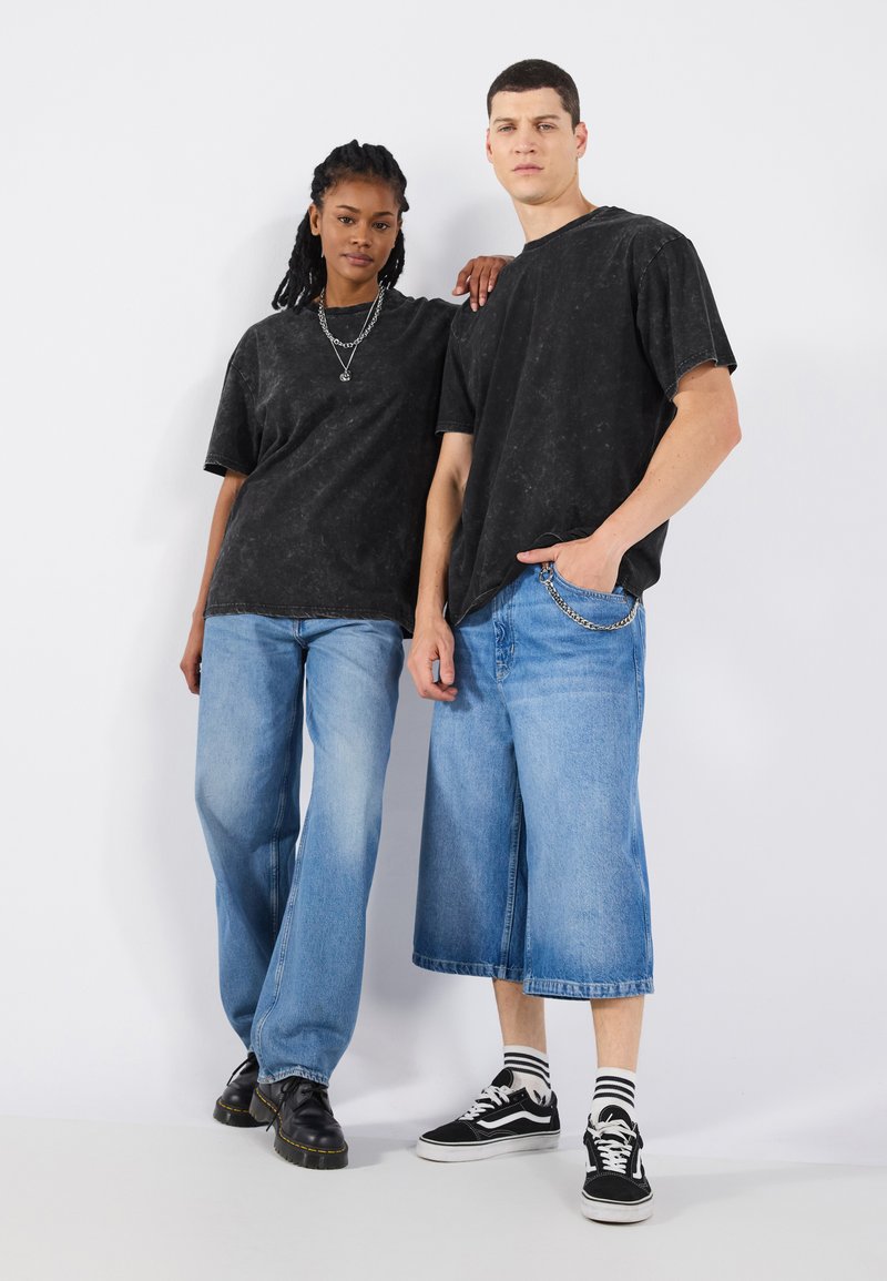 Tricouri supradimensionate cu imprimeu tie-dye negru, asortate cu blugi denim cu picioare largi de culoare albastru deschis. Modelul masculin poartă pantofi negri; modelul feminin poartă adidași.