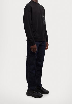 Mand iført sort sweatshirt med frontlomme, mørkeblå jeans og sorte sneakers, stående mod en ensfarvet baggrund.