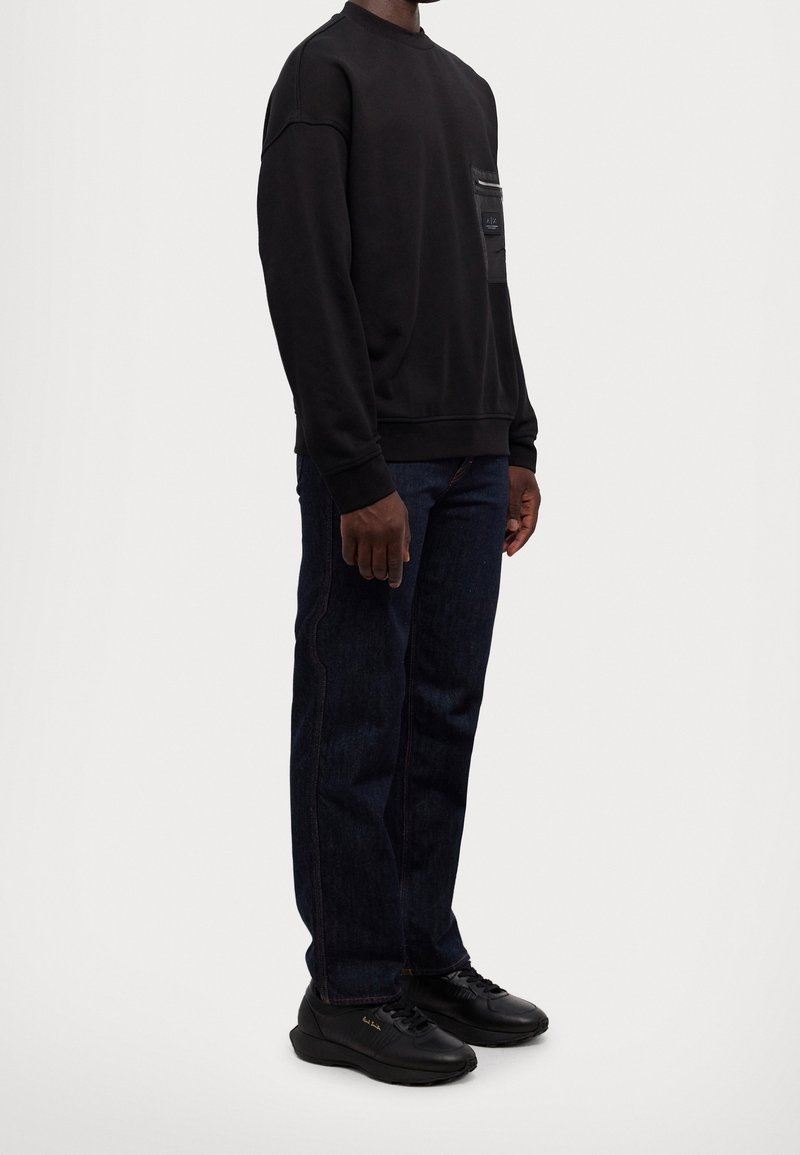 Homme portant un sweat-shirt noir avec poche avant, jean bleu foncé et baskets noires, debout devant un fond uni.
