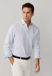 Mann trägt hellblau gestreiftes Hemd mit hochgekrempelten Ärmeln, weiße Hose und dunkelbraunen Gürtel, schaut zur Seite.