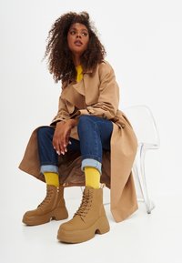 Stivaletti alla caviglia in gomma color tan con tacco spesso, chiusura con lacci e cerniera laterale, abbinati a jeans in denim arrotolati e a un maglione giallo.