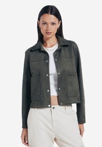 Jasnozielona kurtka z zamszu z kołnierzykiem, zatrzaskami, dwiema przednimi kieszeniami i skróconym krojem, noszona na białej bluzce.