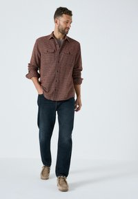 Mees punases tekstuurses särgis, tumesinistes teksades ja tansuede kingades, seisab ühe käega taskus ja vaatab kõrvale.