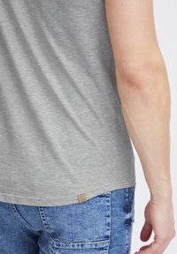 Graues Baumwoll-T-Shirt mit abgerundetem Saum, das ein kleines Logo-Patch nahe dem unteren Ende zeigt. Trägt es mit blauen Jeans.