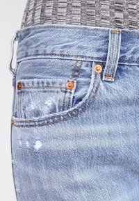 Los jeans de mezclilla azul claro presentan un diseño desgastado con bordes deshilachados y remaches de cobre. La tela muestra una apariencia suave y texturizada.