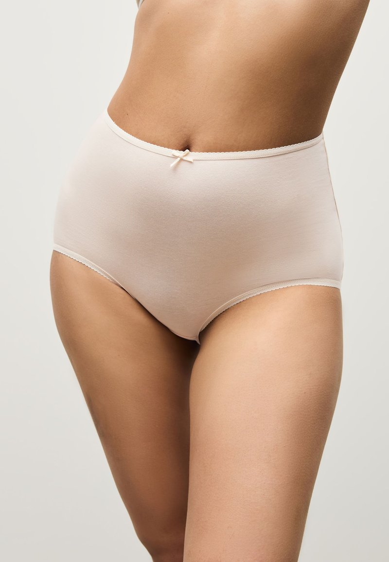 Ropa interior femenina beige de cintura alta con pequeño detalle de lazo, usada en la parte inferior del torso y la parte superior de los muslos de una persona de pie.