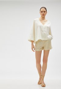 Løst siddende hvidt linnedbluse med V-hals, parret med beige shorts og gyldne sandaler med remme, sat mod en ensfarvet baggrund.