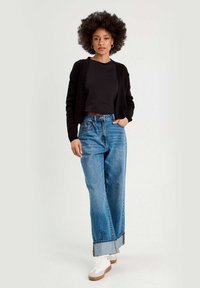 Svart kort cardigan över en svart topp, matchad med ljusblå vida jeans med vikta fållar och vita sneakers.