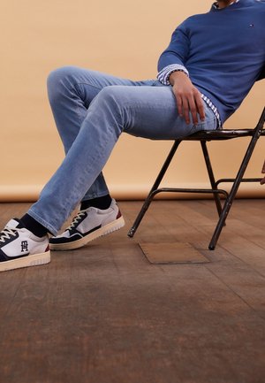 Persona con vaqueros azules, suéter azul y zapatillas sentada en una silla plegable de metal sobre un fondo beige en un suelo de madera.