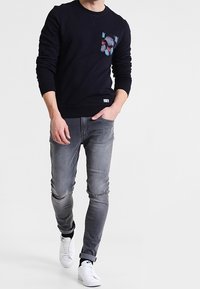 Marineblaues Sweatshirt mit einer Blumenmuster-Tasche, kombiniert mit hellgrauen Skinny Jeans und weißen Sneakers. Lässiger, moderner Look.