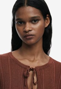 Rustrød ribbet sweater med rund halsudskæring og bindelukning, der har en blød tekstur og subtil glans. Nærbillede af stofdetaljen.