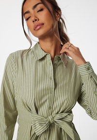 Groen en wit gestreept knoopshirt met een strik in de taille. Kenmerkt zich door een kraag en lange mouwen met knoopmanchetten.