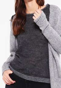 Femme portant un pull ras du cou gris foncé sous un gilet en tricot gris clair, avec une main ajustant le col du gilet.