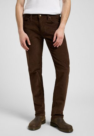 Man draagt donkerbruine, nauwsluitende ribfluwelen broek met bruine leren laarzen en een wit overhemd, staand tegen een effen lichte achtergrond.